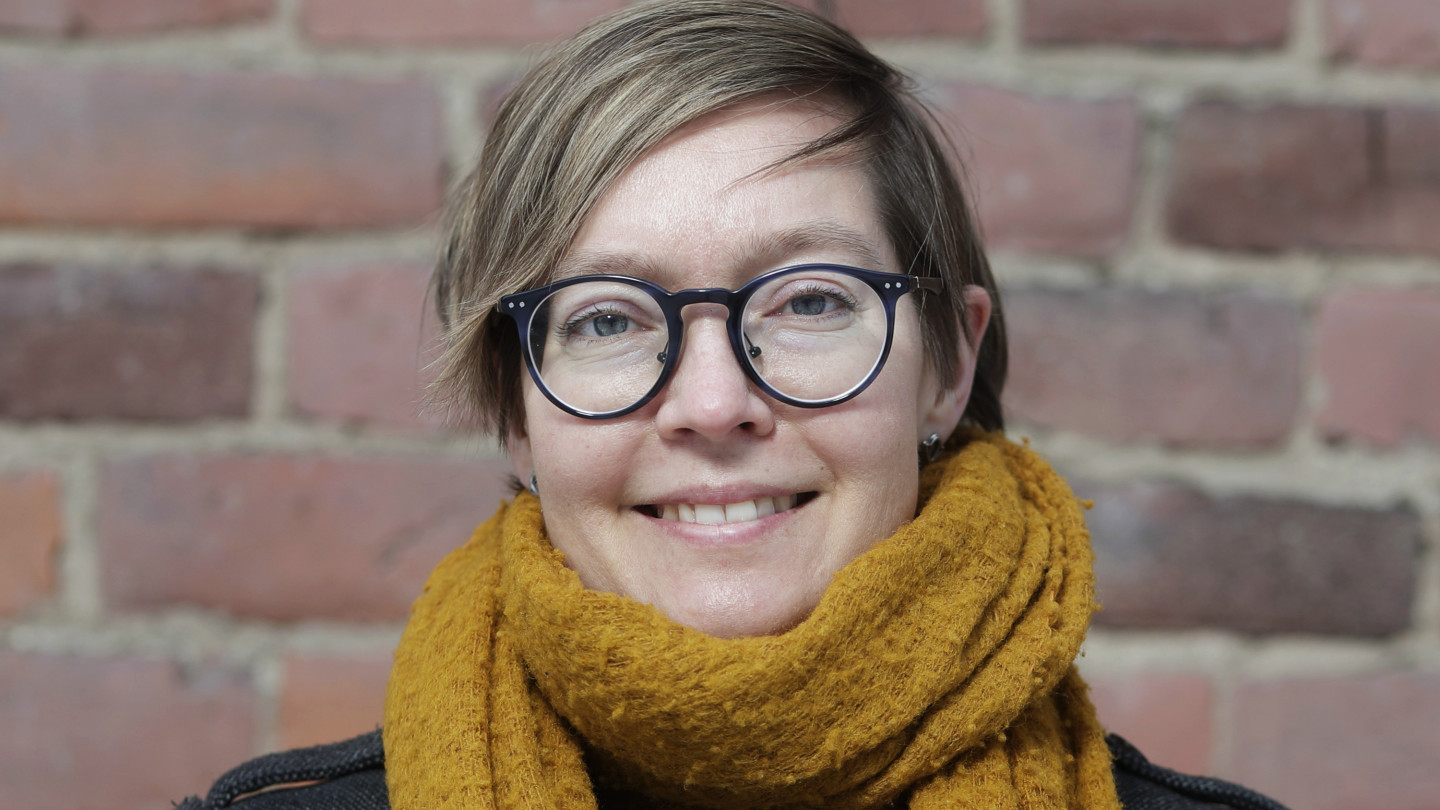 Eerika Koskinen-Koivisto punaisen tiiliseinän edessä. Hänellä on lyhyet ruskeat hiukset ja silmälasit. Kaulassaan hänellä on kirkkaan oranssi kaulahuivi.