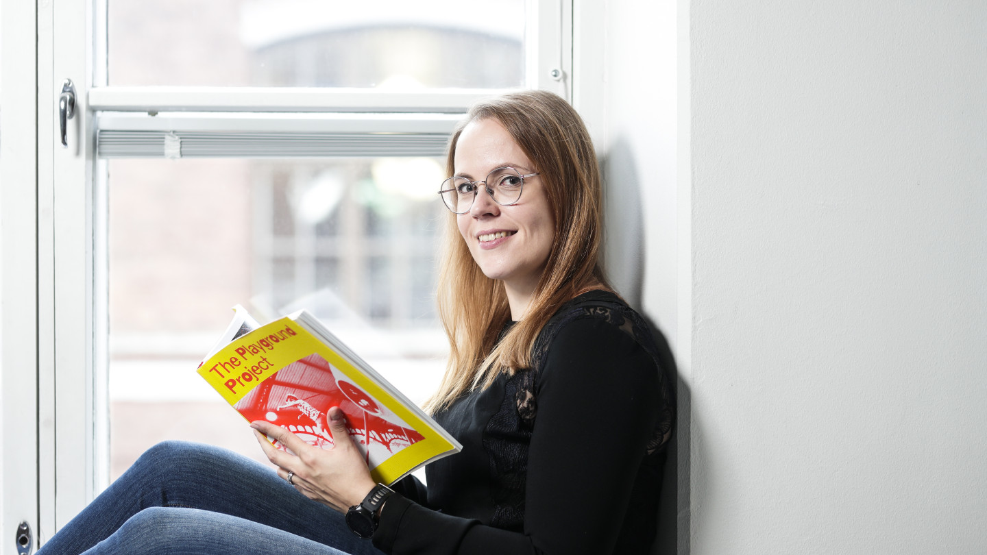 Essi Jouhki istuu suurella ikkunalaudalla lukemassa kirjaa. Hänellä on ruskeat pitkät hiukset, musta paita ja siniset farkut.
