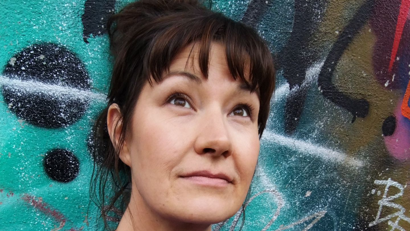 Satu Hotari, jolla on tumman ruskeat puolipitkät hiukset ponnarilla. Hän seisoo graffitin edessä.