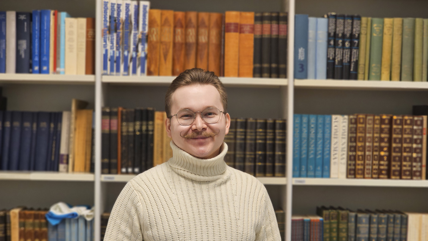 Heikki, jolla on tummanruskeat lyhyet hiukset, silmälasit ja yllään beige villapaita seisoo hymyillen kirjahyllyn edessä.