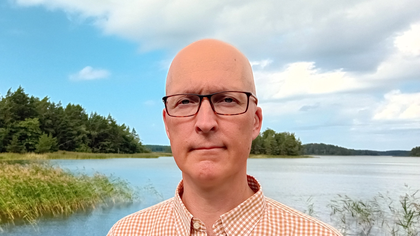 Väitöskirjatutkija Mikko Taipale. Hän katsoo kameraan, taustallaan kaislikkoinen järvimaisema ja pilvinen taivas.