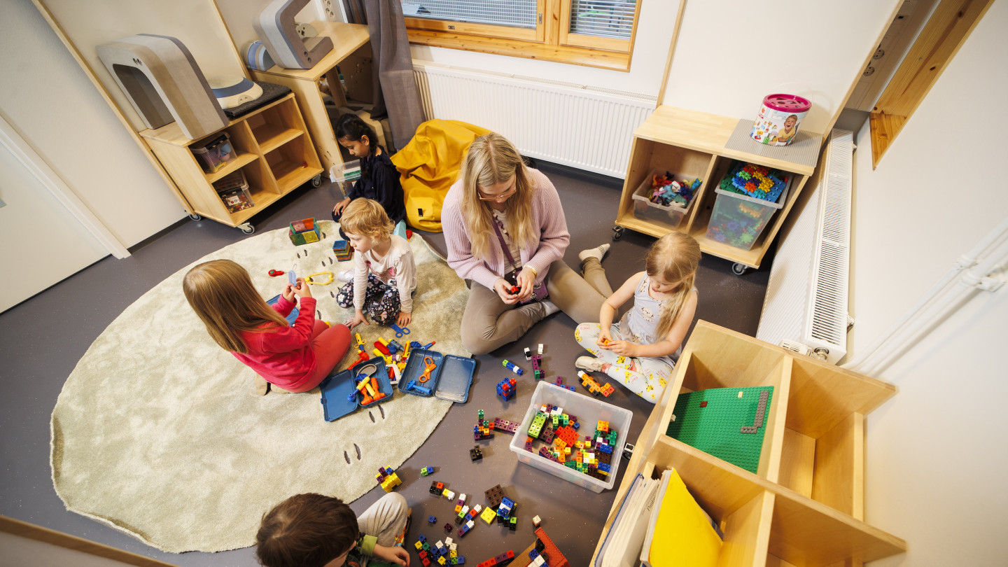 Varhaiskasvatuksen opettaja ja lapset istuvat lattialla leikkimässä legoilla.