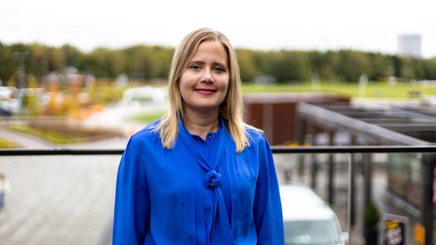 Elisa seisoo ulkona ja hymyilee kameralle. Puolikuva. Hänellä on sininen paita.