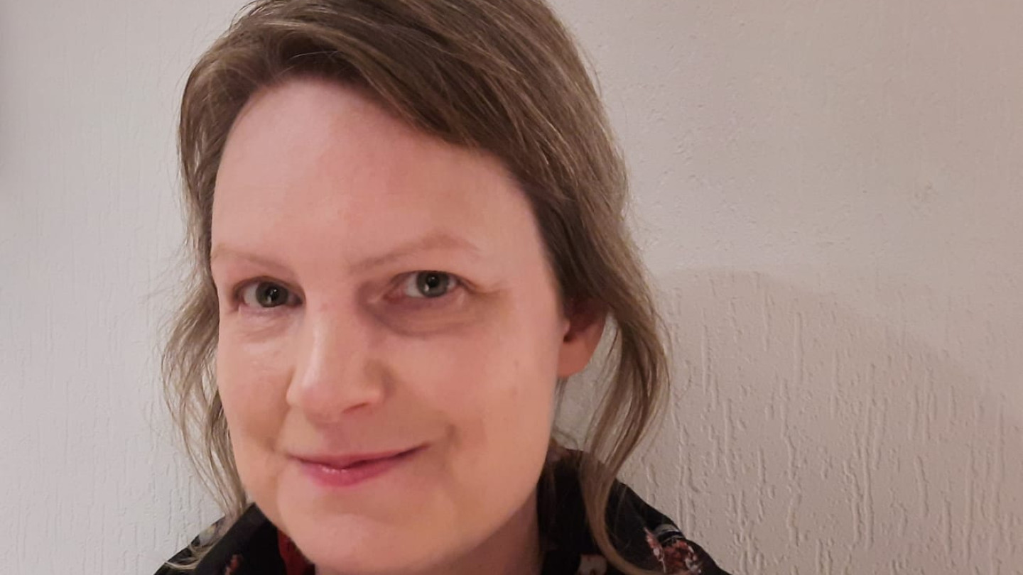Sanna Dziomba katsoo kameraa hymyillen. Hänellä on musta paita ja musta, kukallinen huivi kaulassaan. Tausta on valkoinen.