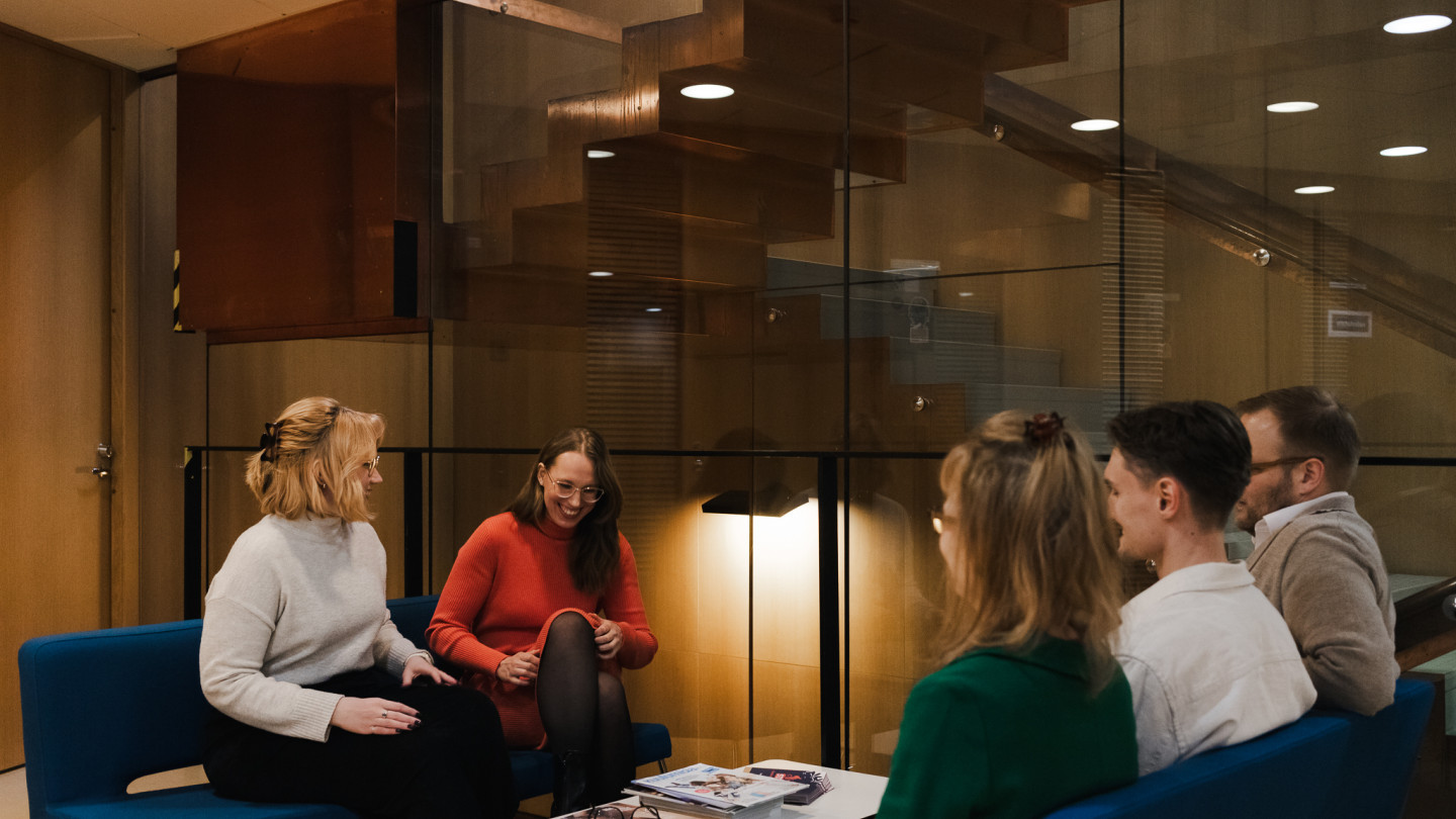Viisi ihmistä istuvat vastakkain kahdella eri sohvalla ja keskustelevat. Taustalla lasiseinä ja sen takana näkyy portaikko ylös.