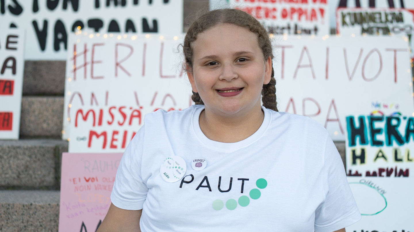 Elokuvan päähenkilö Aida istuu Eduskuntatalon portailla ja hymyilee. Aidalla on päällään valkoinen Tpaita, jossa on Autismiyhdistys PAUTin logo, sekä vaaleanvihreät shortsit, joissa on valkoisia kuvioita, jotka muistuttavat laineita. Aidan taakse portaille on aseteltu kantaa ottavia kylttejä. 