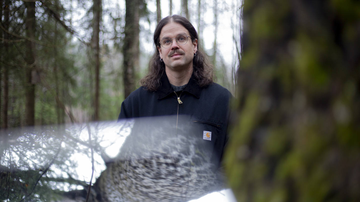 Ate Tervonen seisoo metsässä puun vieressä. Ate Tervonen in a forest, standing next to a tree.