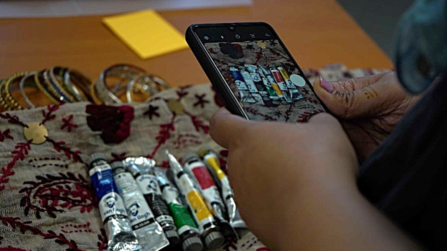 A photograph of tubes of oil paints on a decorative cloth. The photo also shows the hands of someone taking a photograph of the paints on a mobile phone.