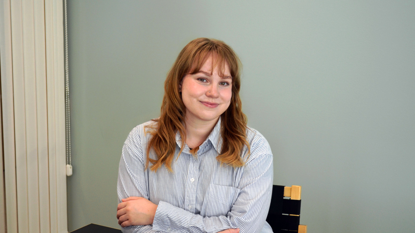 EmmiJulia katsoo kameraan hymyillen. Hän istuu penkillä ja nojaa pöytään. 