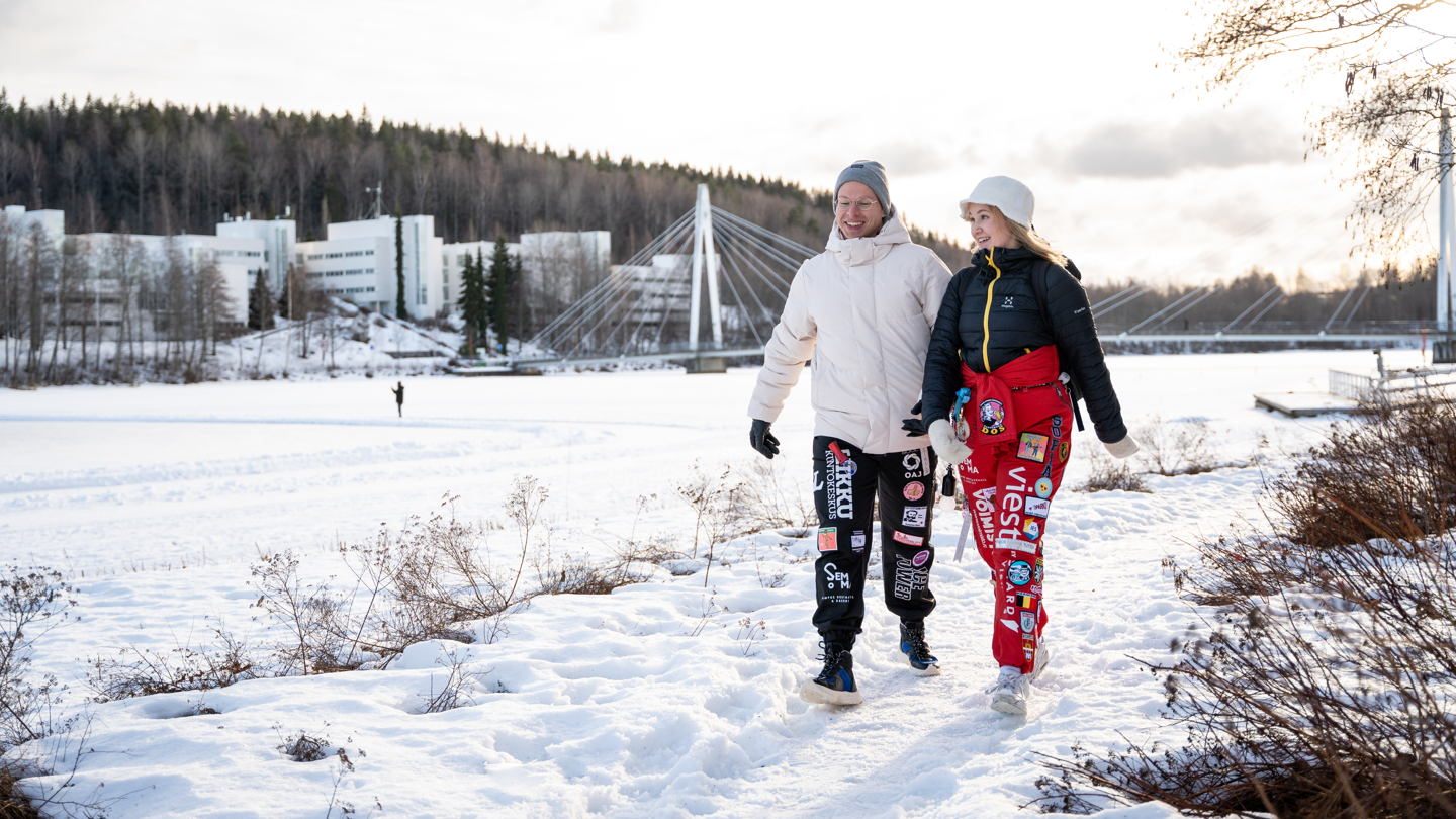 Kaksi opiskelijaa kävelee talvisessa maisemassa, taustalla Ylistön silta ja kampus. 