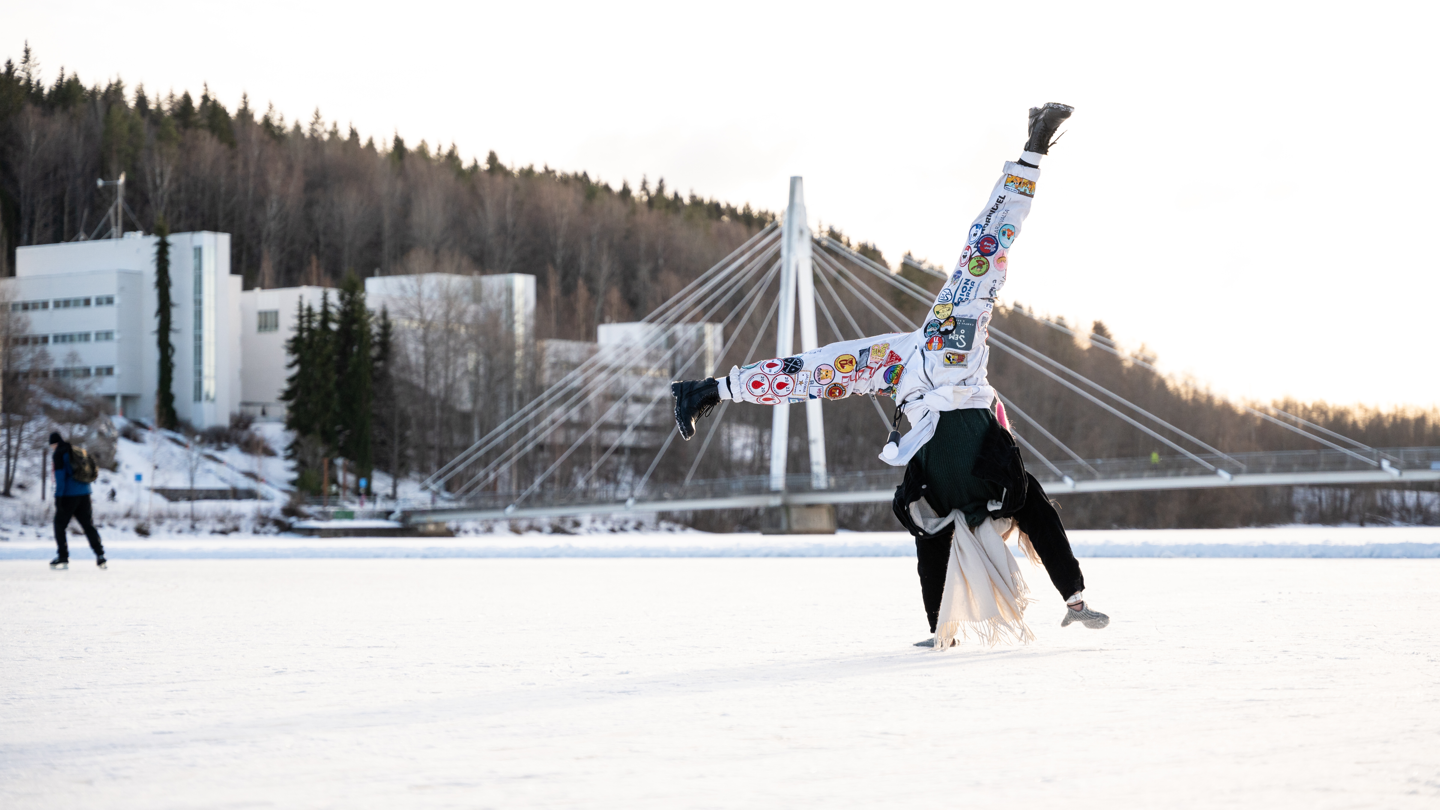 Opiskelija tekee kärrynpyörän järven jäällä, taustalla Ylistön silta ja kampusta. 