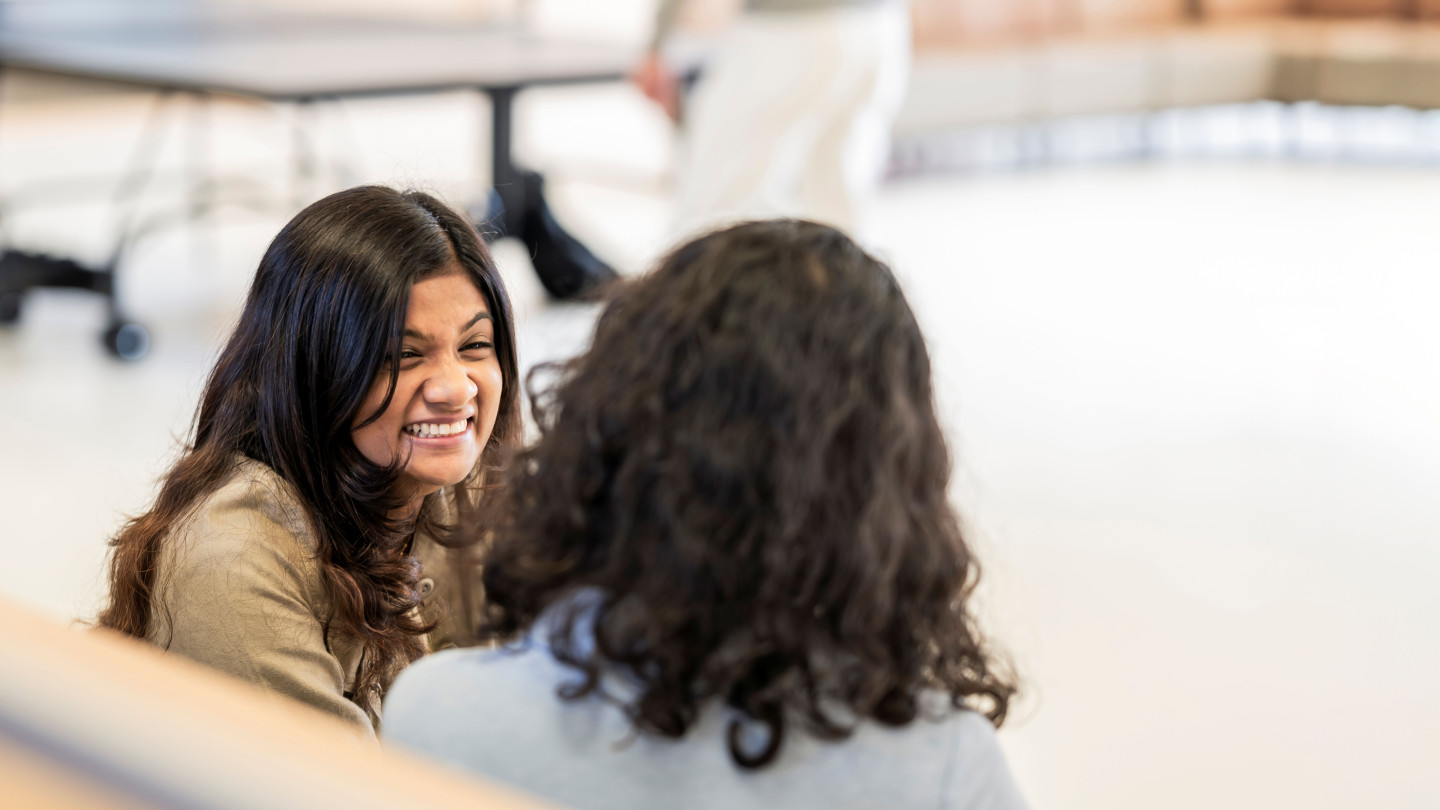 Two students sitting indoors on a campus, talking together.