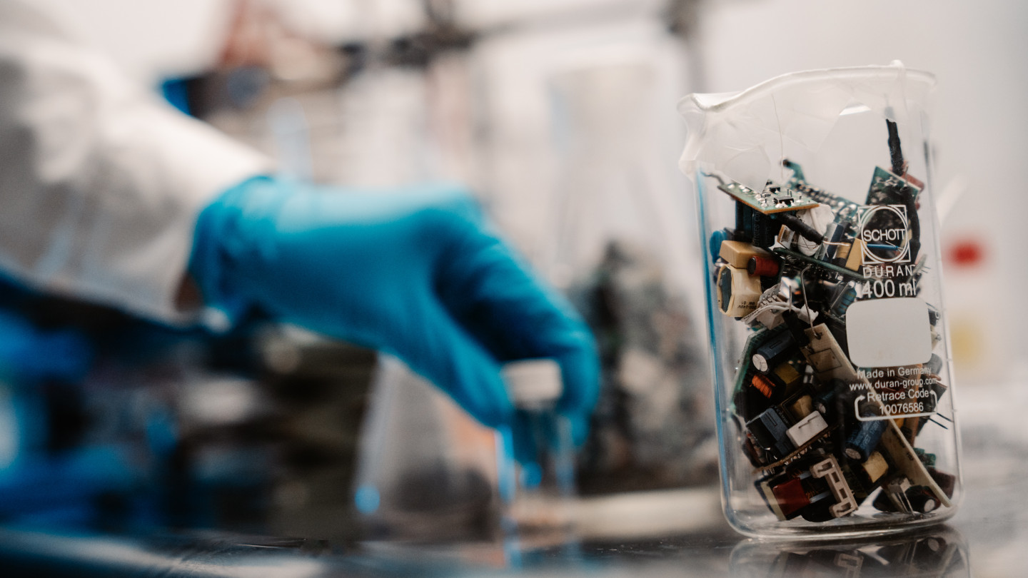 Electronic waste in a see-through beaker and laboratory personnel moving samples in the background 