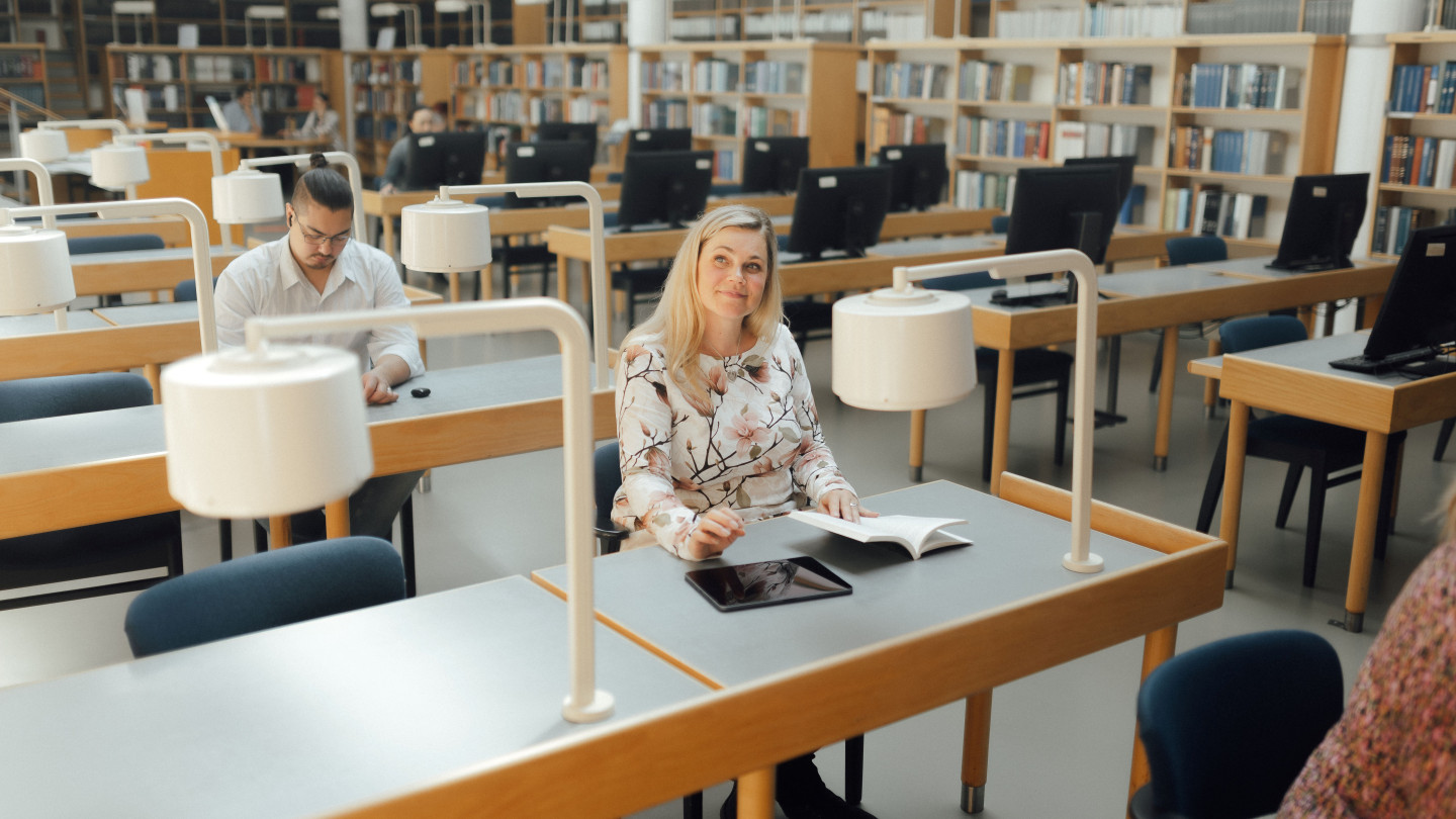Kuvassa opiskelee aikuisopiskelijoita kirjastossa. Nainen opiskelee kirjan parissa mietteliään näköisenä..