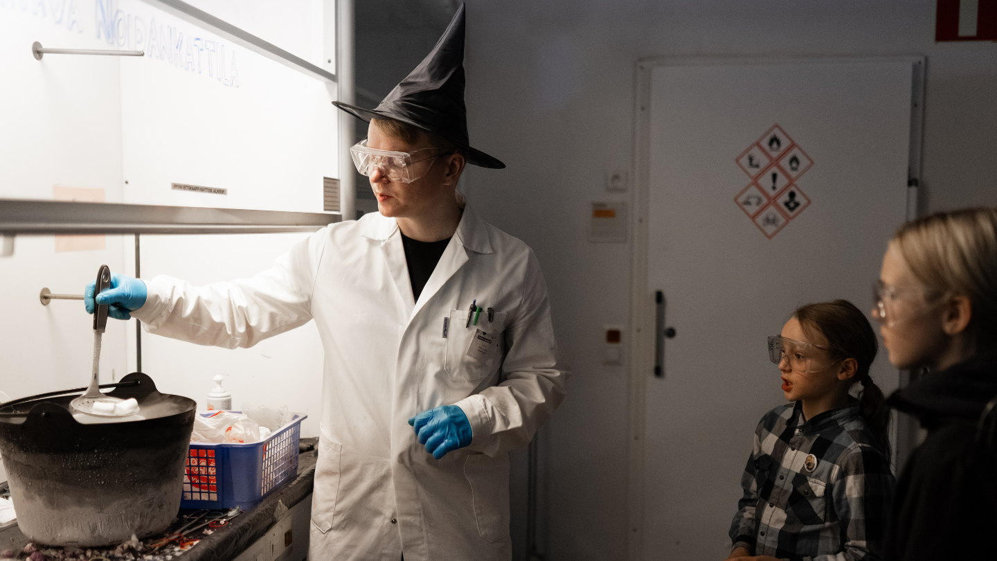 Tutkija, jolla on yllään laboratoriotakki ja noitahattu, nostaa metallilusikalla jotain harmaasta kattilasta.