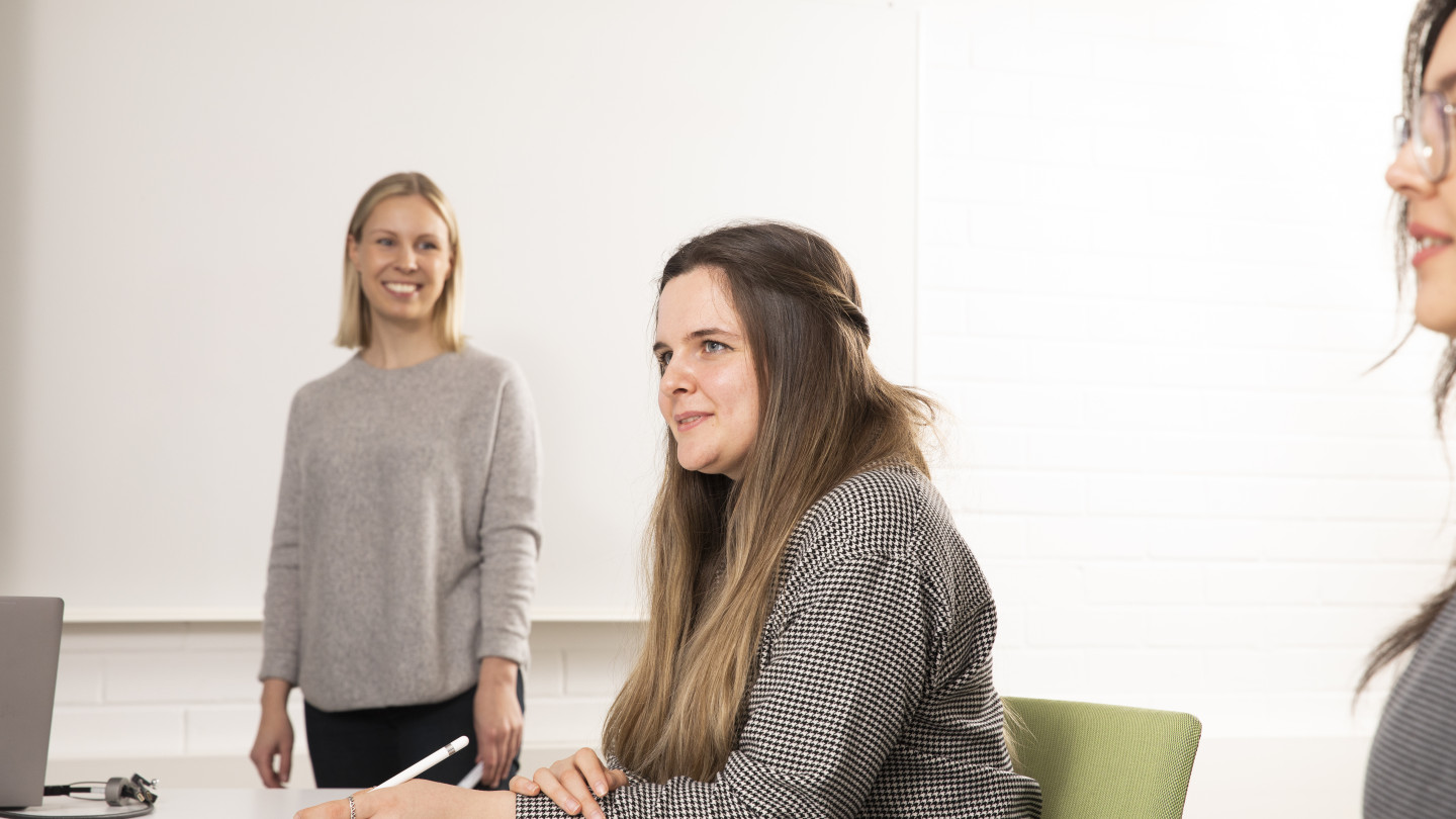 Kaksi naista istuvat pöydän ääressä kuuntelemassa ja yksi nainen kuuntelee seisten. Naiset ovat opiskelemassa .