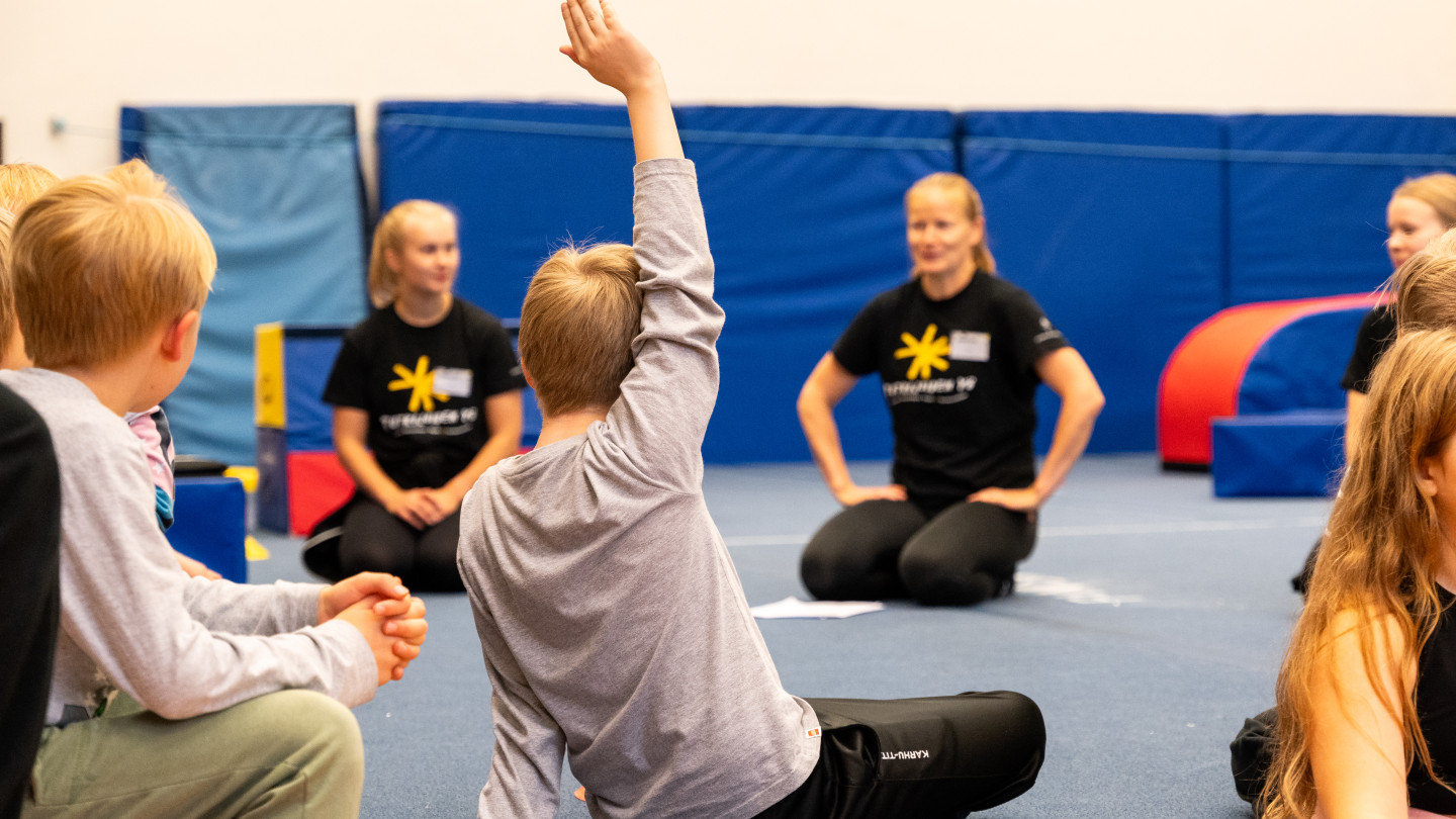 Kaksi aikuista ja lapsia kerääntyneenä istumaan jumppasalin lattialle Tutkijoiden yö-tapahtumassa. Harmaapaitainen lapsi viittaa.