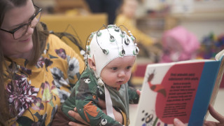 Vauva katselee kirjan sivua äitinsä sylissä istuen. Vauva osallistuu tutkimukseen, ja hänen päässään on aivotoimintaa rekisteröivin anturein varustettu myssy.