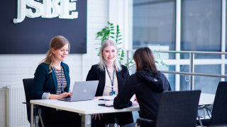 Kolme henkilöä työskentelee työpöydän ääressä, JSBEkyltti