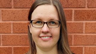 Miia Kuha, jolla on silmälasit ja ruskeat olkapäille ulottuvat hiukset. Yllään hänellä on musta villatakki ja vaaleansininen paita.