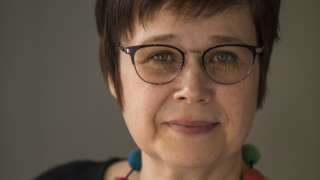 Merja Karjalainen, jolla on lyhyet ruskeat hiukset ja silmälasit. Yllään hänellä on musta paita ja värikäs huovutetuista helmistä tehty kaulakoru.