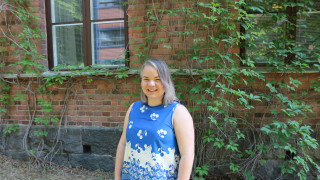 Karoliina hymyilee punatiilisen Historican edessä kesällä. Hänellä on yllään sininen hihaton paita.