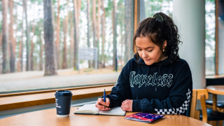 Opiskelija istuu ikkunan ääressä ja tekee merkintöjä vihkoon.