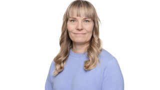 Katri katsoo kameraan hymyillen, hänellä on vaaleat hiukset ja vaaleansininen neulepaita. 
