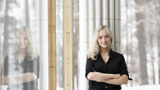 Henkilö katsoo hymyillen ja kädet ristissä puuskassa kameraan. Musta paita ja vaaleat hiukset.