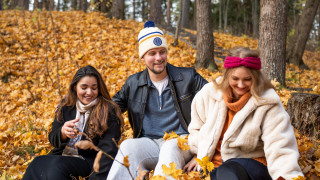 Three students sitting outdoors in an autumn setting, surrounded by colorful fallen leaves.