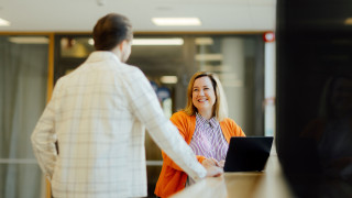 Kaksi opiskelijaa juttelemassa toisilleen sisätiloissa, toinen tietokoneen ääressä.