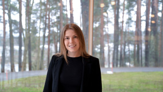 Gratista seisoo mäntymaiseman edessä. Hän on pukeutunut mustaan.
