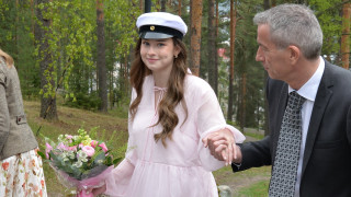 Kuvassa yleinen seppeleensitojatar on kiipeämässä portaita katsoen kameraan. Hänellä on vaaleanpunainen mekko, päässään ylioppilaslakki ja kädessä kukkakimppu.