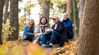Neljä opiskelijaa istuu maassa puiden keskellä syksyisessä maisemassa.