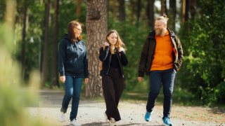 Kolme henkilö kävelee jutellen puistomaisessa, vihreässä ympäristössä
