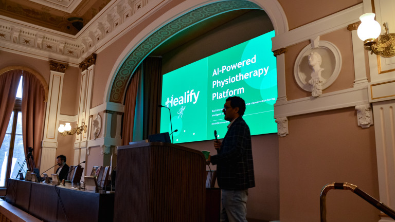 A man presenting in front of a bight green reading "Healify  AIpowered physiotherapy platform". He is standing on a podium and holding a mic.