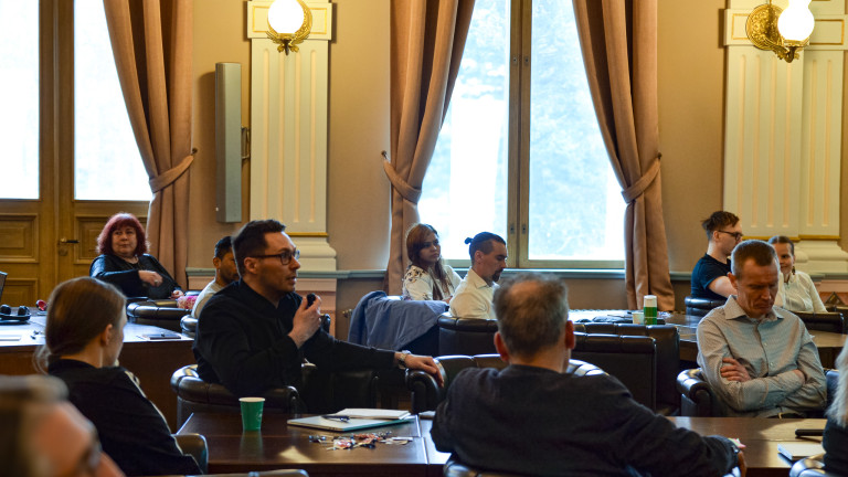 Audience sitting in the Jyväskylä City Hall's Great Hall. A man with a black collar shirt speaking to a microphone in the middle of the photo.