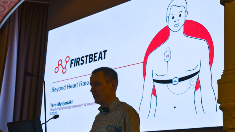 Reading on a big screen "Firstbeat  Beyond Heart Rate" and "Tero Myllymäki, Head of Physiology research and analytics". A man wearing a white collar shirt standing in front of the screen.