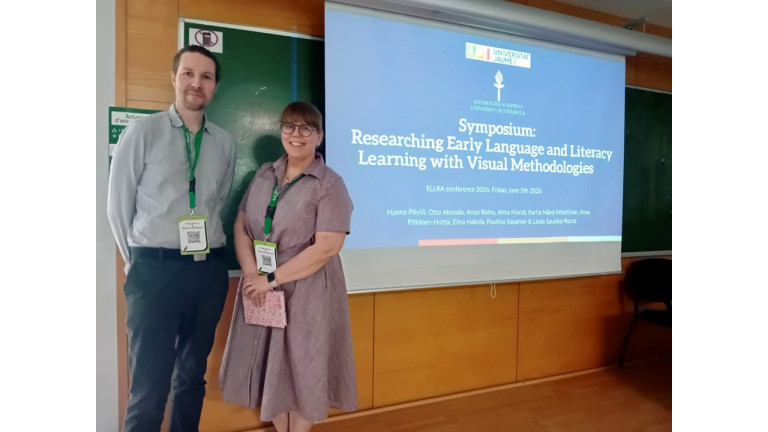 Mies ja nainen seisovat vierekkäin ja hymyilevät kameralle. Molemmilla on kaulassa konferenssin nimikyltit. Heidän vieressään valkokankaalle heijastettu otsikkodia, jossa lukee Symposium: Researching Early Language and Literacy learning with Visual Methodologies.