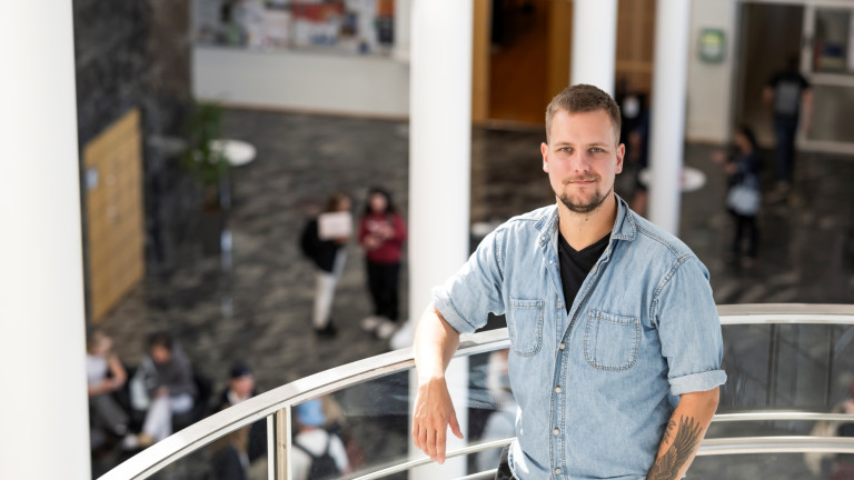Opiskelija katsoo kameraan ja nojaa portaiden kaiteeseen, taustalla muita ihmisiä. 