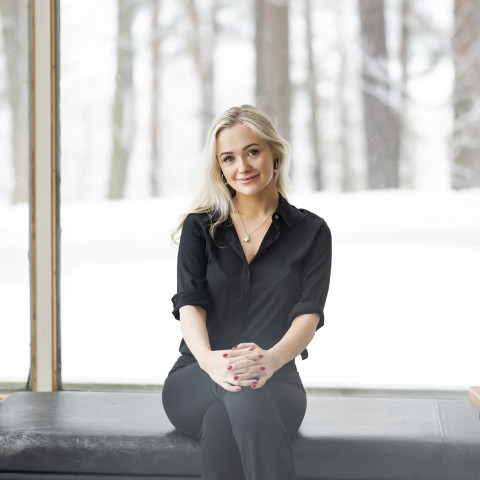 Henkilö istuu jakkaralla jalat ristissä. Mustat housut ja musta paita. Vaalea tukka auki. 