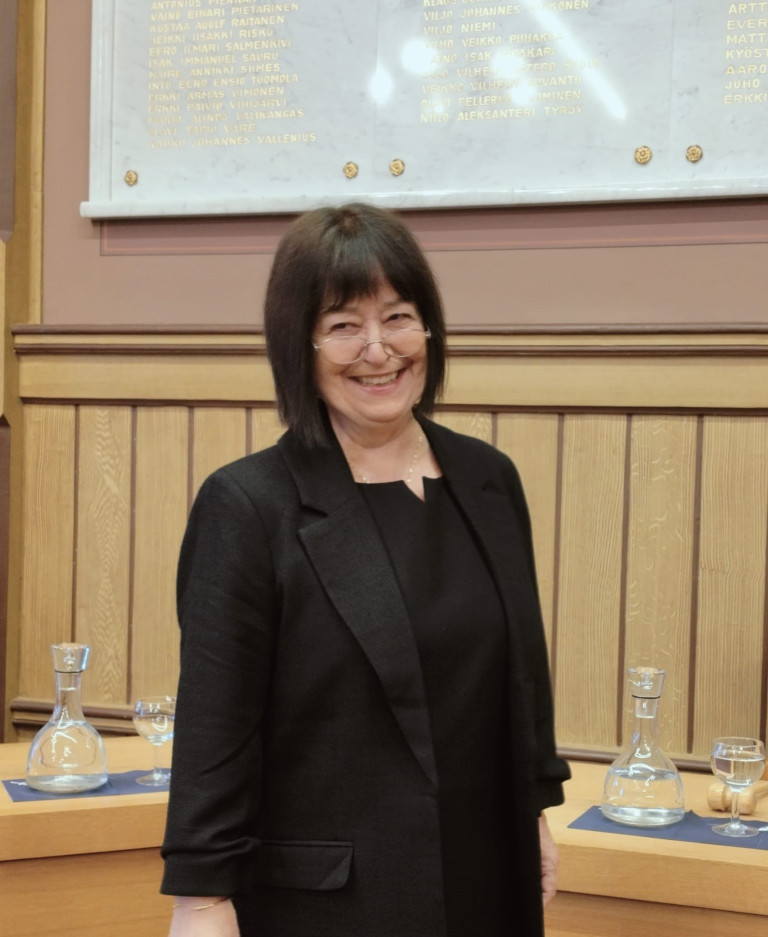 Henkilö seisoo ja hymyilee hieman vasemmalle ohi kameran. Päällä musta jakku ja paita. Tummanruskeat polkkahiukset ja silmälasit.