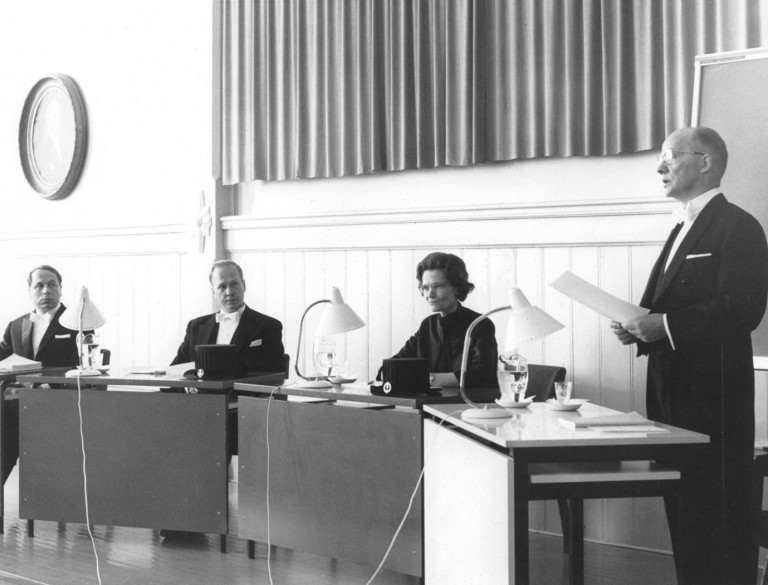 Professori Faina Jyrkilä väitöstilaisuuden kustoksena yliopiston vanhassa juhlasalissa vuonna 1969. Muut kuvan henkilöt ovat vastaväittelijät Juhani Karvonen (vas.) ja Kalevi Heinilä sekä väittelijä Uuno Tuomola. Kuva: Jyväskylän yliopiston tiedemuseo.